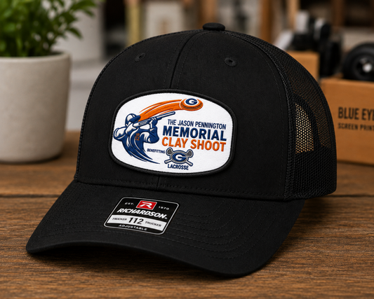 A black Richardson 112 trucker hat with a white, blue, and orange 'The Jason Pennington Memorial Clay Shoot' patch on the front, displayed on a wooden table with a blurred background.