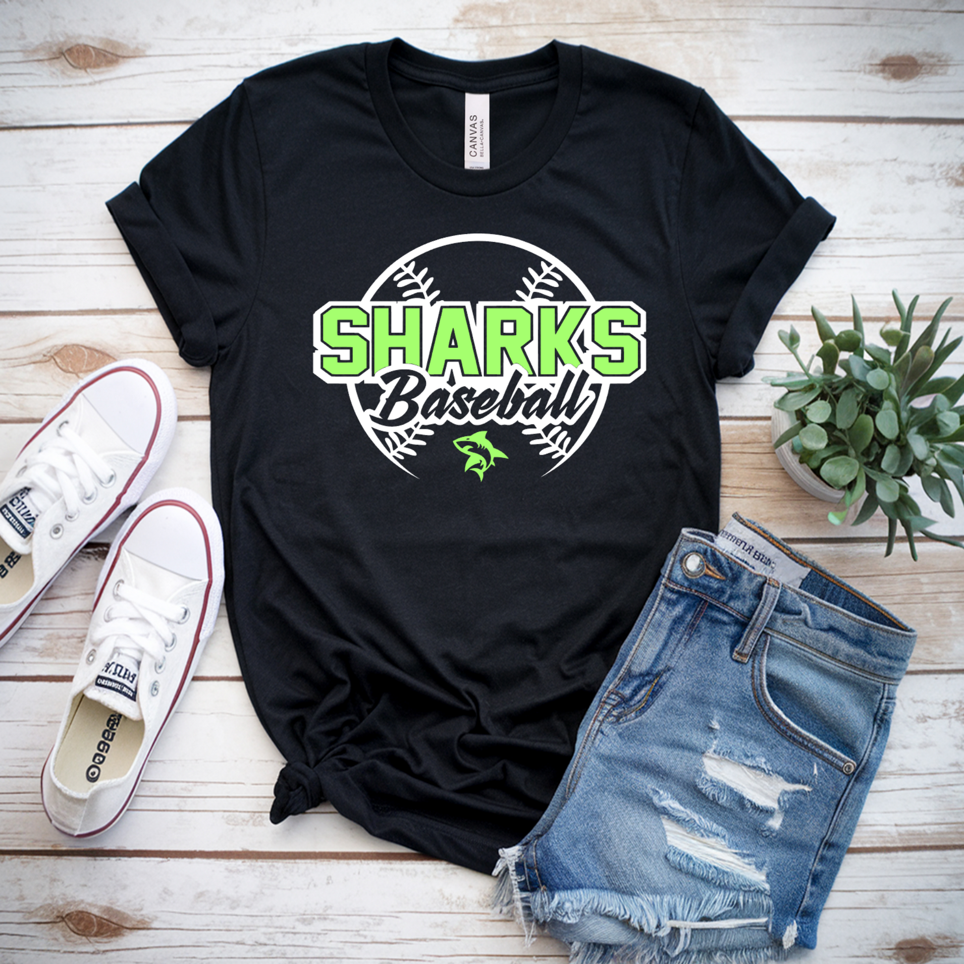 Black t-shirt with 'Sharks Baseball' design, white sneakers, denim shorts, and a plant on a wooden surface.