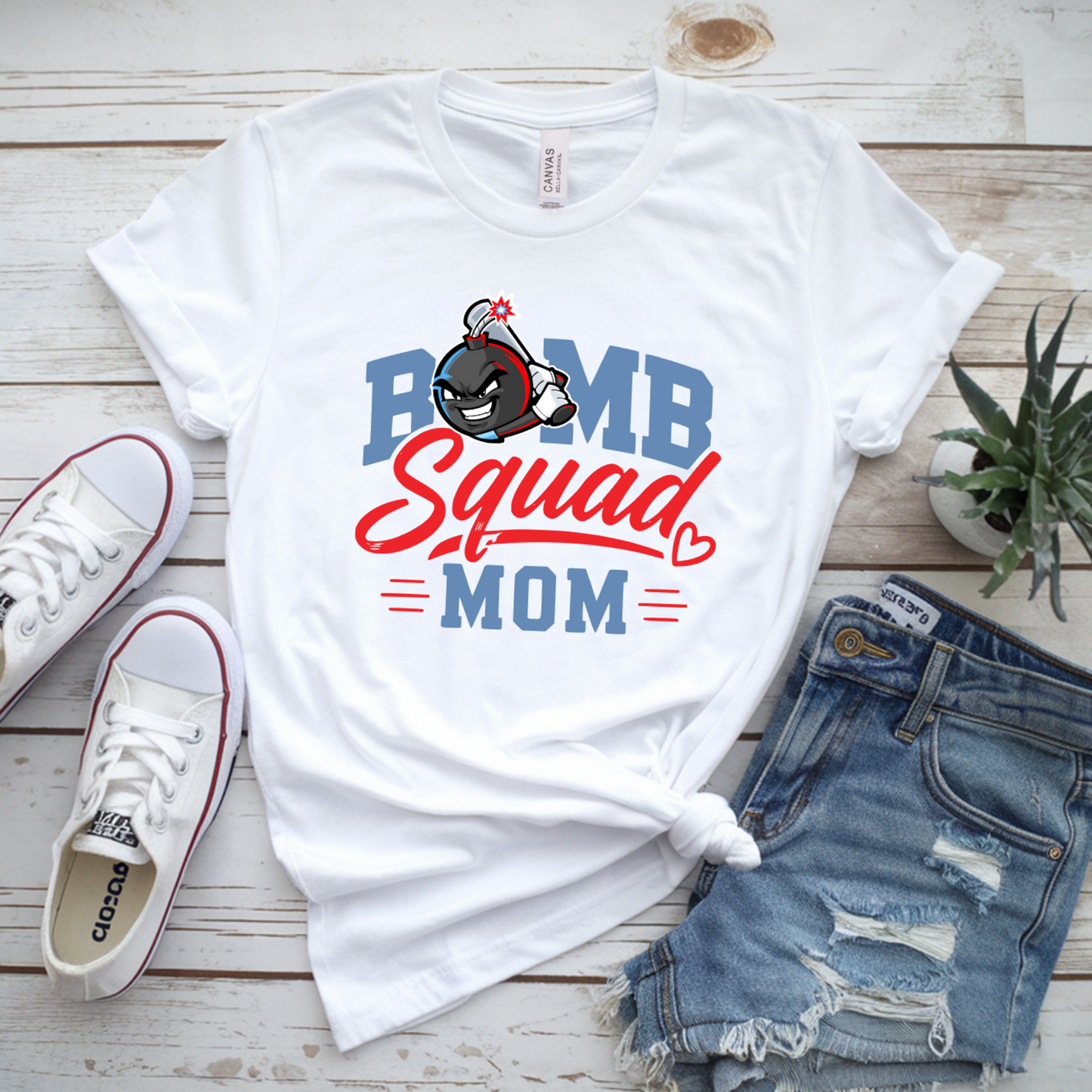White graphic T-shirt laid flat on a whitewashed wood surface, styled with white Converse sneakers, distressed denim shorts, and a small potted succulent. The shirt graphic reads "PUMP Squad MOM" in blue and red lettering with a cartoon character above it.