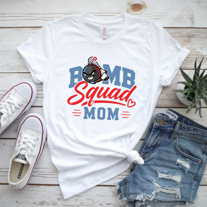 White graphic T-shirt laid flat on a whitewashed wood surface, styled with white Converse sneakers, distressed denim shorts, and a small potted succulent. The shirt graphic reads "PUMP Squad MOM" in blue and red lettering with a cartoon character above it.