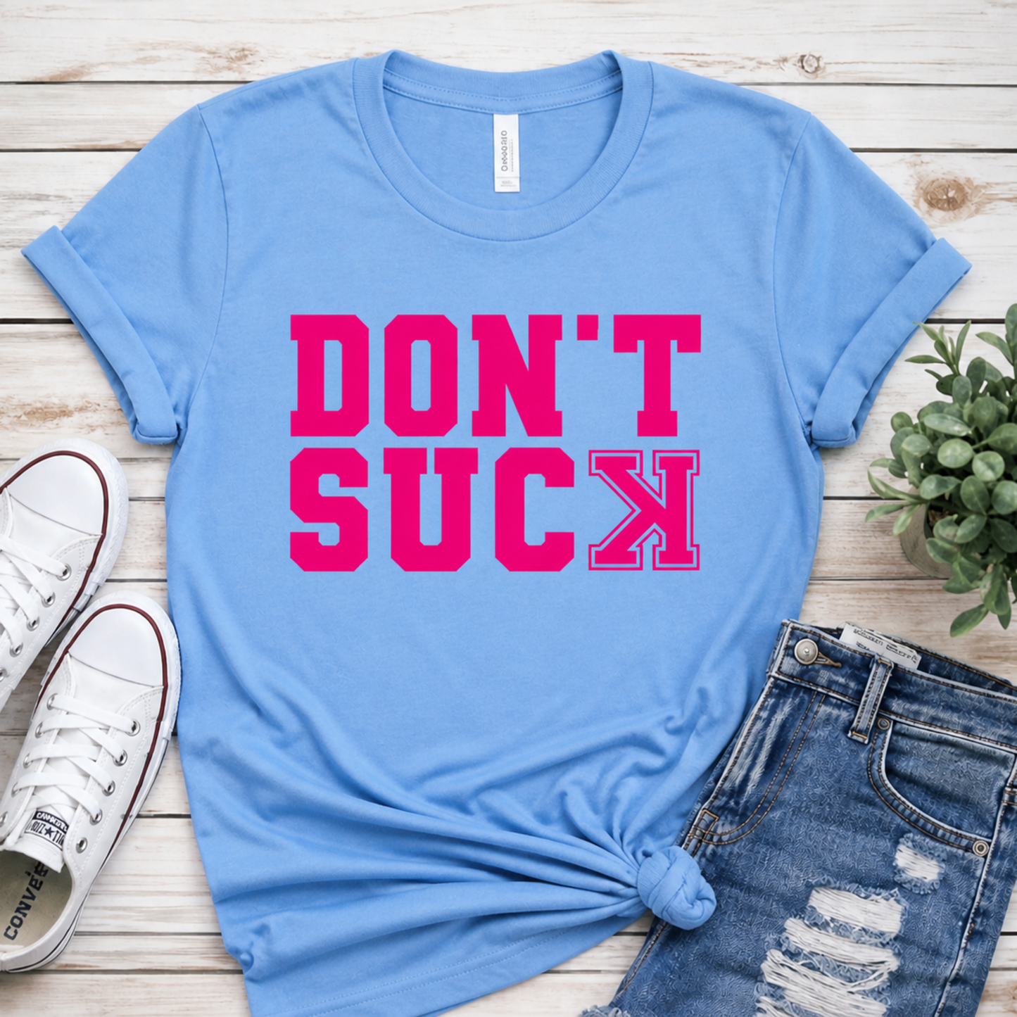 Blue t-shirt with 'DON'T SUCK' in pink on a wooden surface with shoes and jeans.