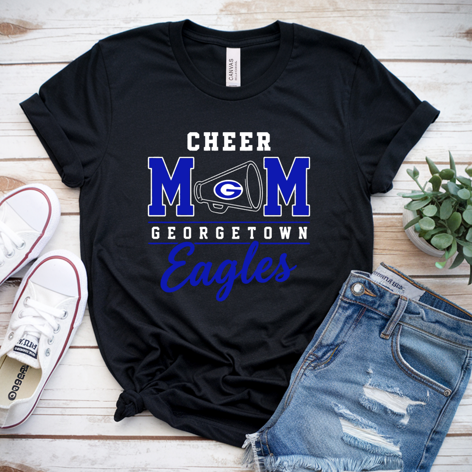 A black graphic T-shirt laid flat on a white wood background, styled with white sneakers, distressed denim shorts, and a small potted plant. The shirt reads “CHEER MOM GEORGETOWN Eagles” with a large blue and white Georgia 'M' graphic.