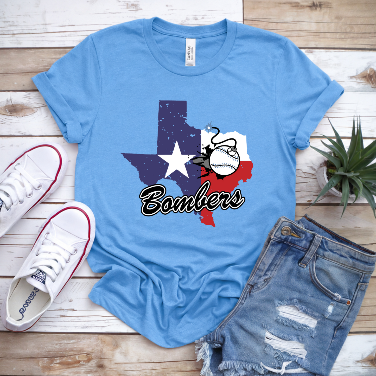 Blue t-shirt with 'Bombers' and Texas map design, paired with white sneakers and denim shorts on a wooden surface.
