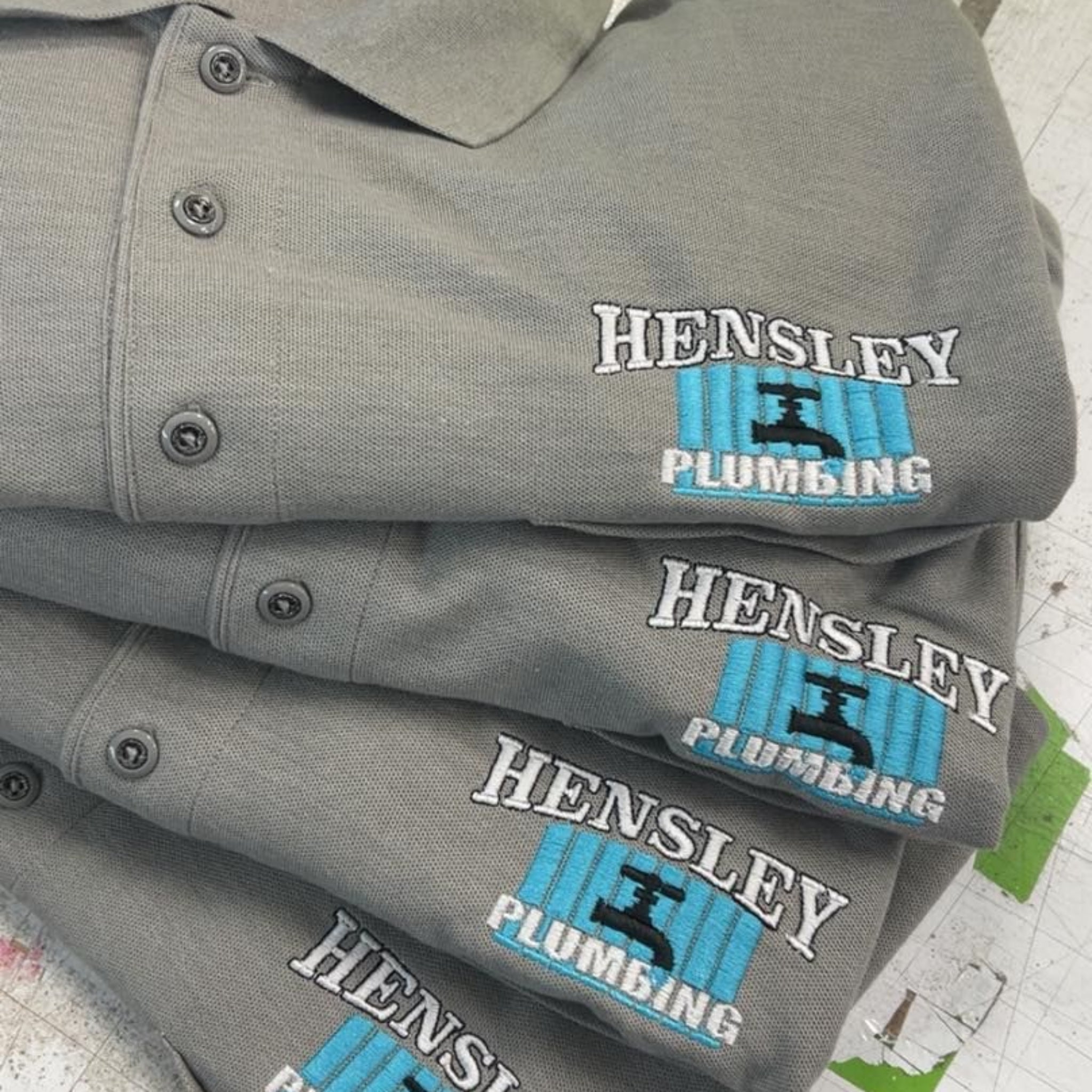 Stack of gray shirts with 'Hensley Plumbing' logo on a newspaper background