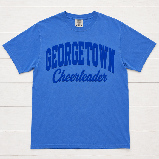 A royal blue short-sleeve t-shirt with 'GEORGETOWN Cheerleader' printed in dark blue across the front, laid flat on a white wood plank background.