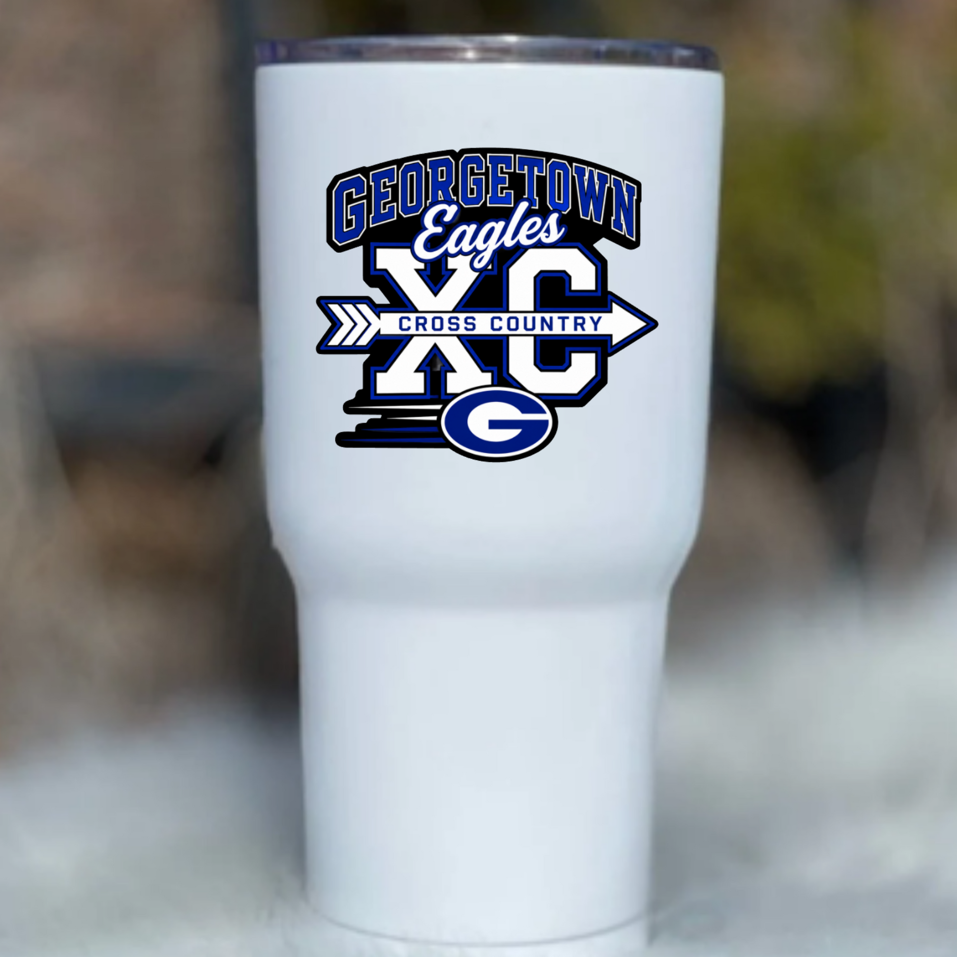 A white insulated tumbler with a clear lid, featuring a blue and white 'Georgetown Eagles XC Cross Country' graphic on the front, set against a blurred outdoor background.