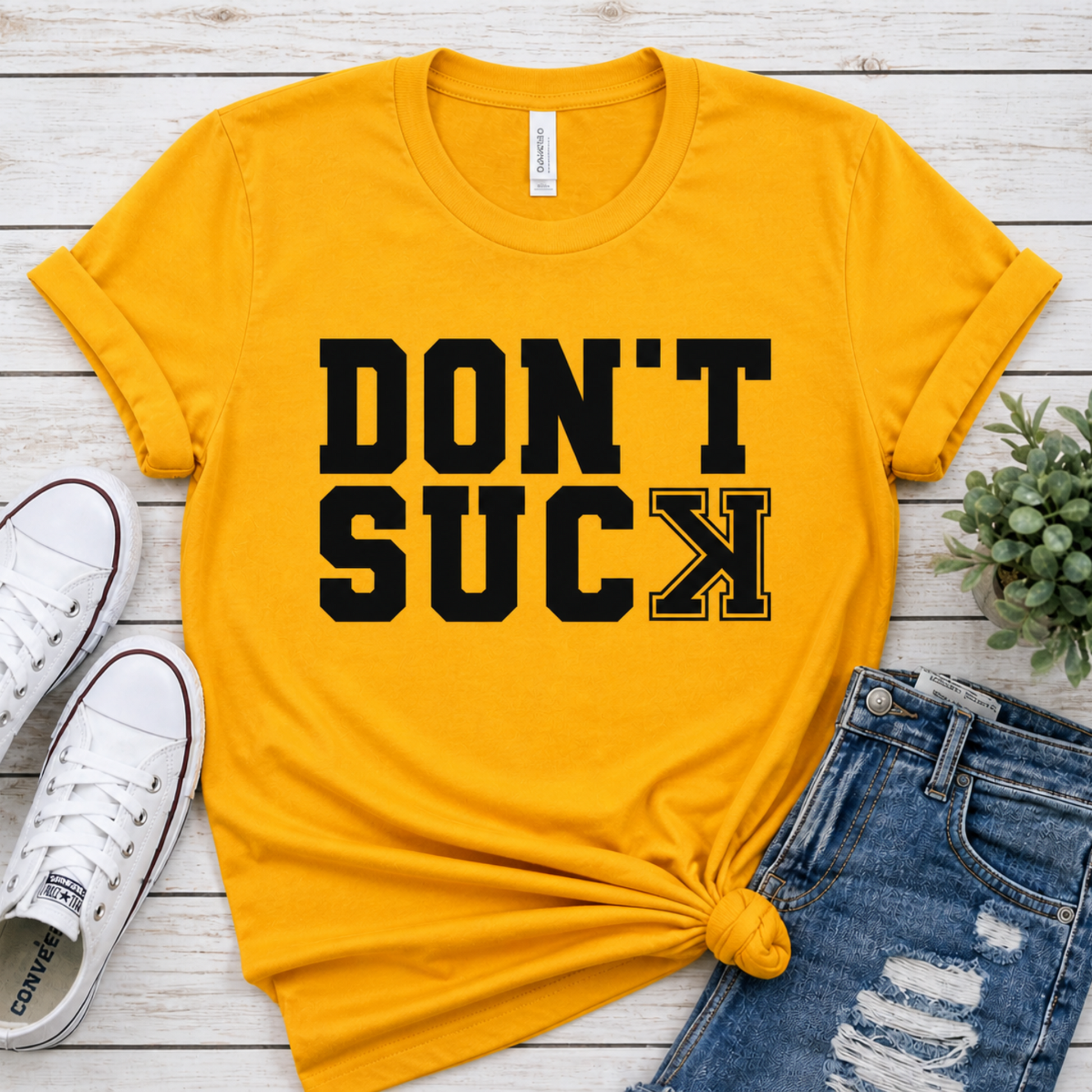 Yellow t-shirt with 'DON'T SUCK' text, white sneakers, and jeans on a wooden surface.