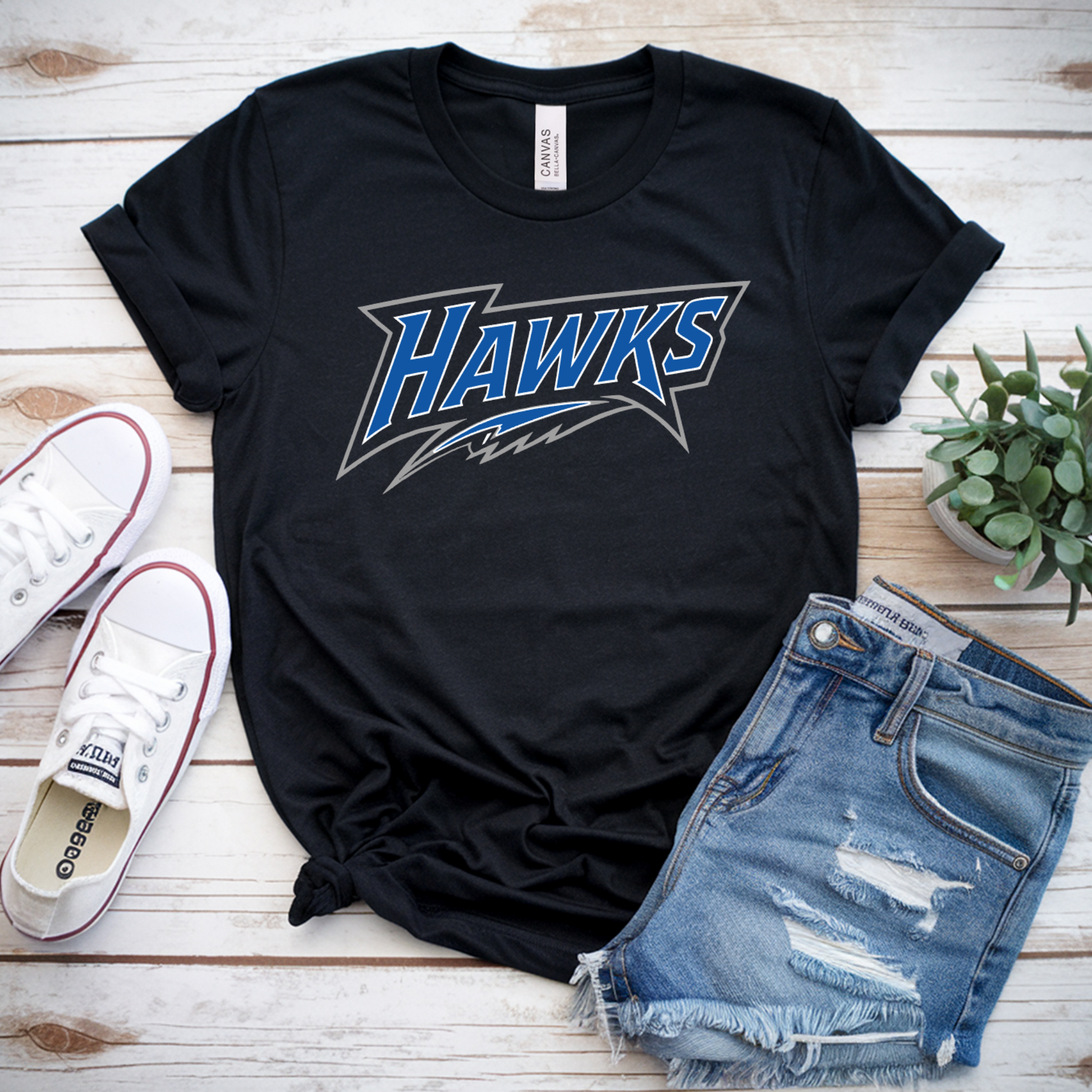 A black graphic t-shirt with 'HAWKS' in blue and white lettering, styled in a flat lay with distressed denim shorts, white canvas sneakers, and a small potted succulent on a white wood plank background.