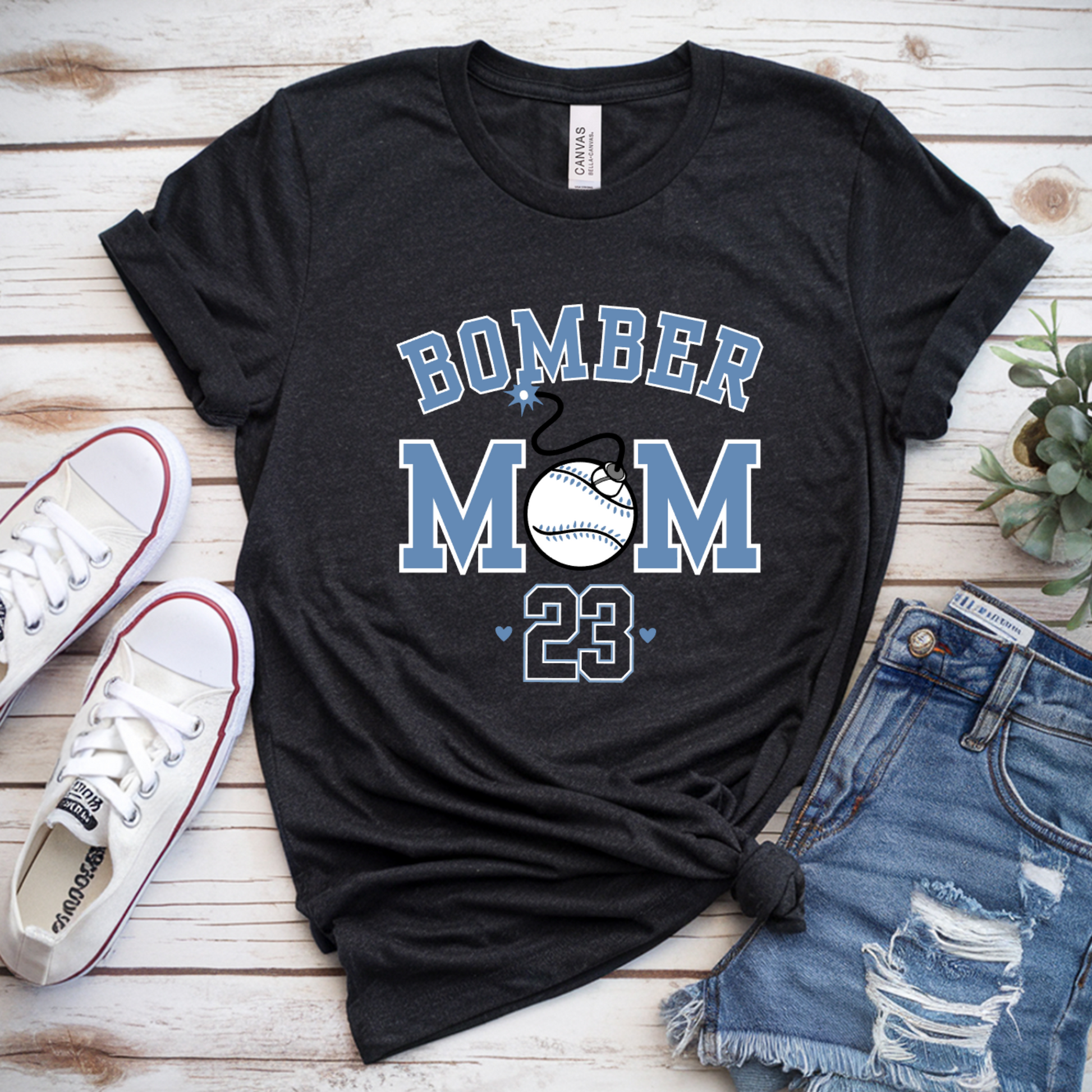 Black t-shirt with 'Bomber Mom 23' text, white sneakers, denim shorts, and a plant on a wooden surface.