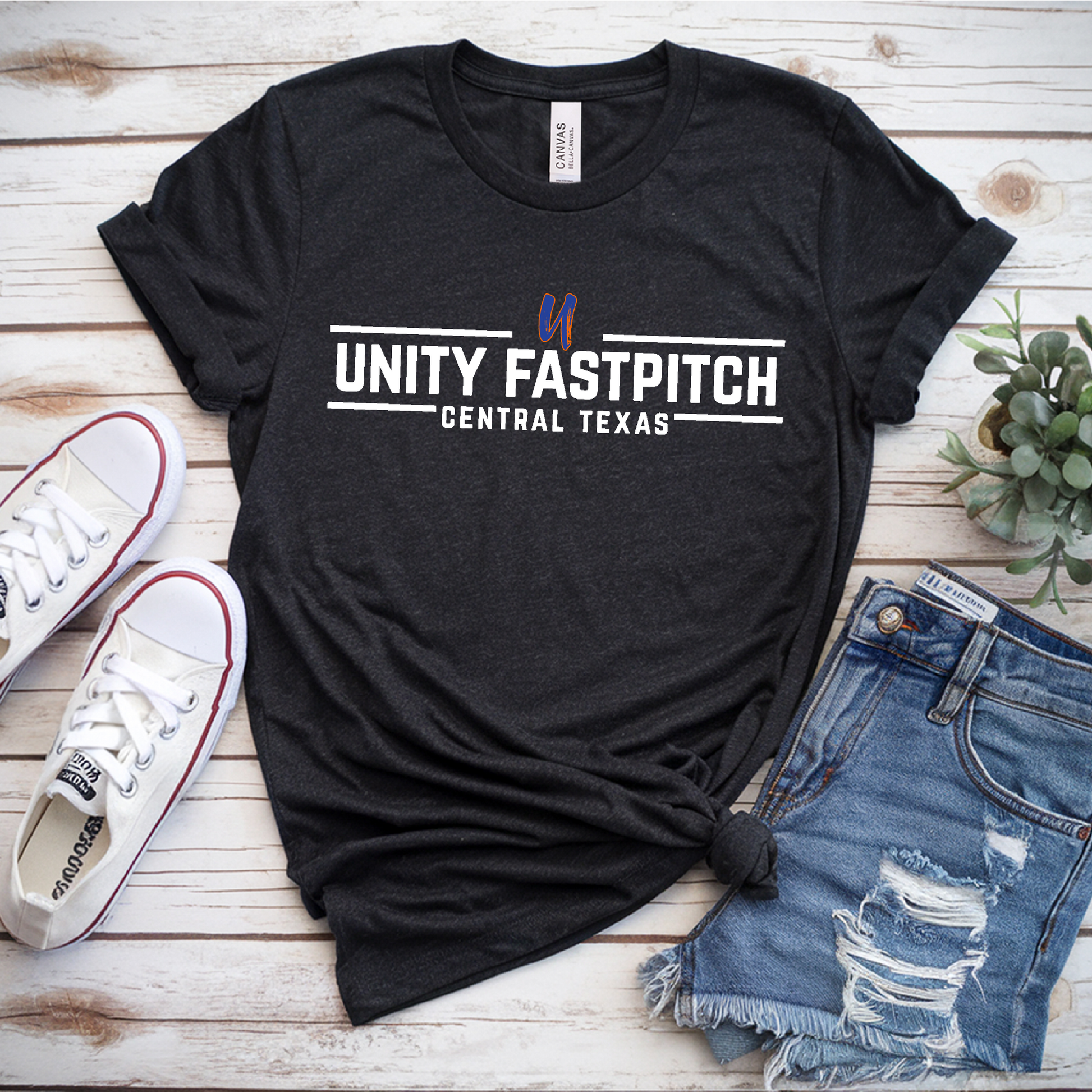 Black t-shirt with 'Unity Fastpitch Central Texas' text, white sneakers, and denim shorts on a wooden surface.