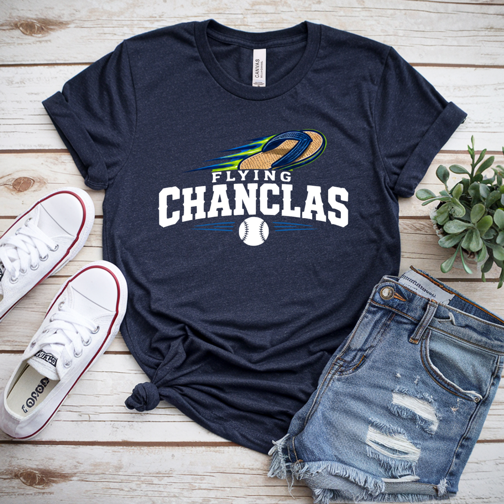 Navy t-shirt with 'Flying Chanclas' design, white sneakers, denim shorts, and a plant on a wooden surface.