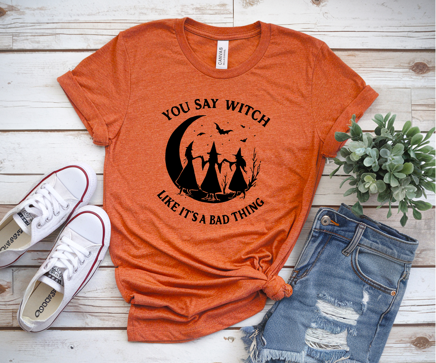 Orange t-shirt with Halloween graphic and text, paired with white sneakers and ripped jeans on a wooden surface.