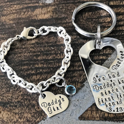 Silver bracelet with 'Daddy's Girl' charm and keychain with engraved text on a wooden surface.