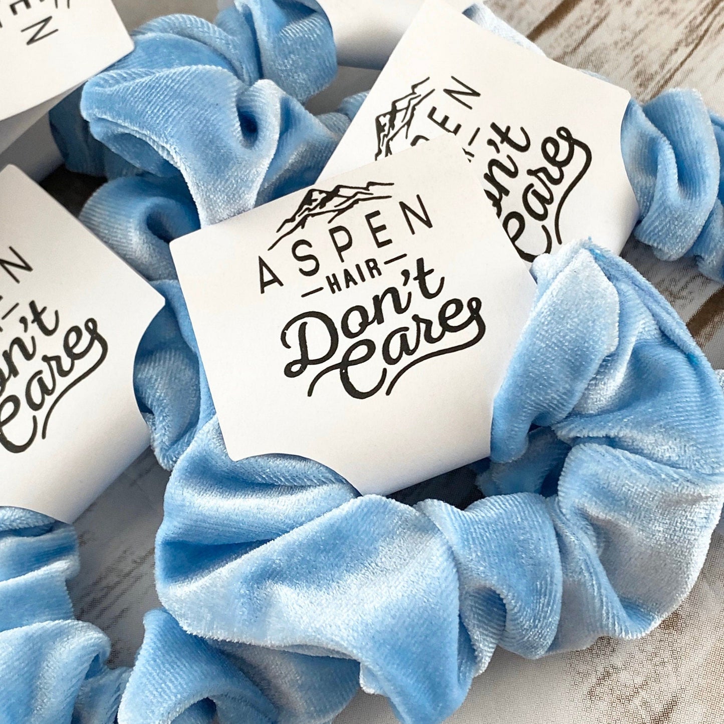 Blue scrunchies with 'Aspen Hair Don't Care' labels on a wooden surface