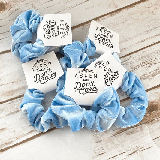 Three blue scrunchies with white labels on a wooden surface