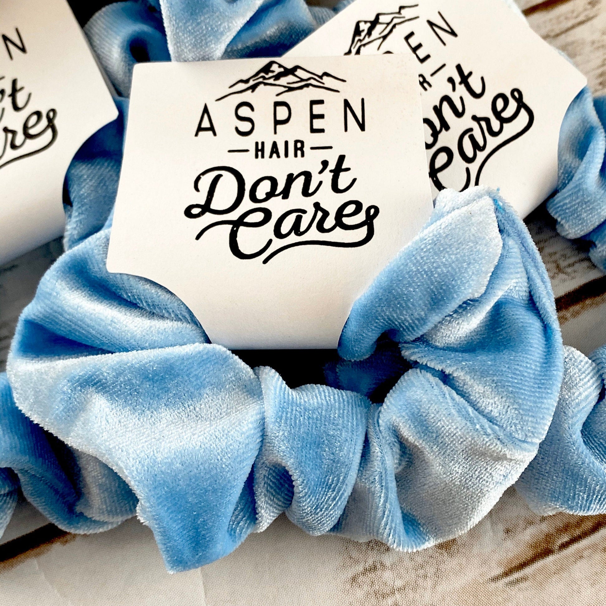 Blue scrunchie with 'Aspen Hair Don't Care' text on a wooden surface