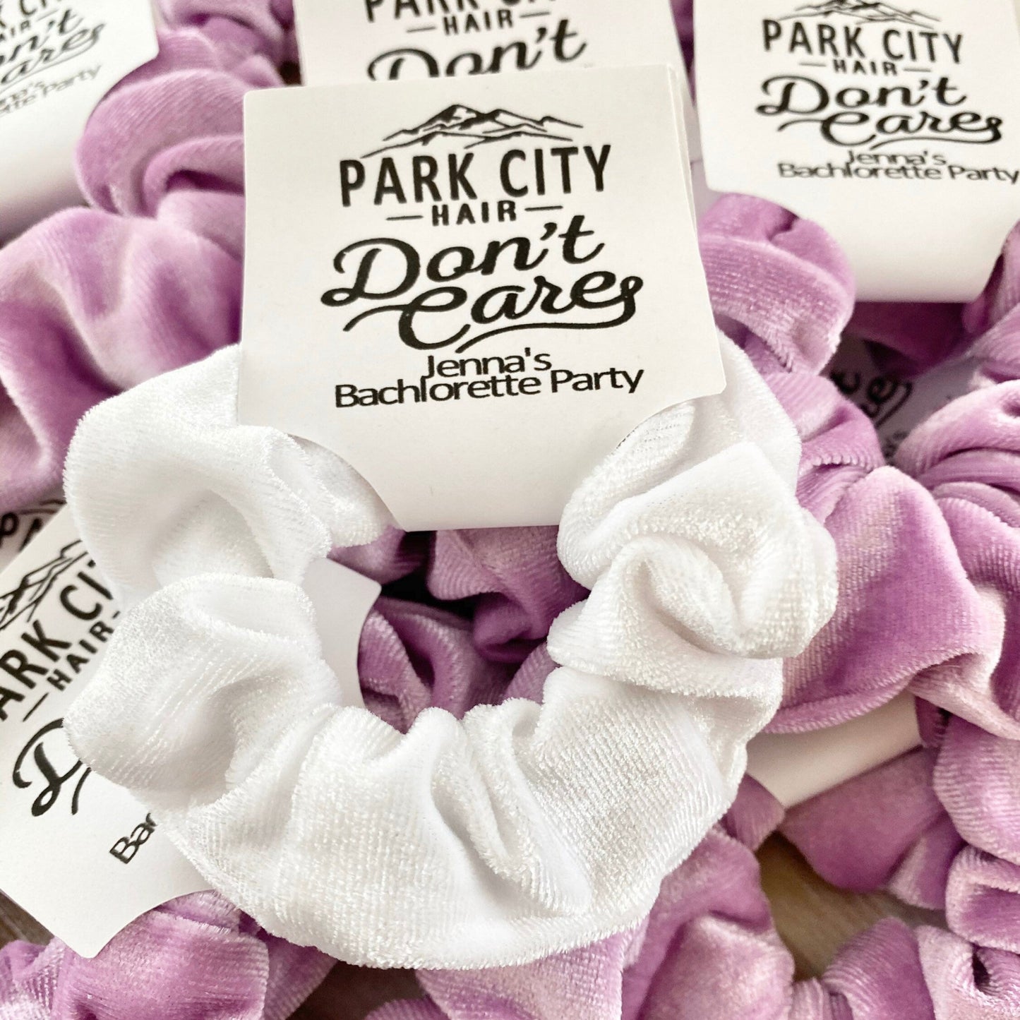 White and purple scrunchies with 'Park City Hair Don't Care' labels on a wooden surface.