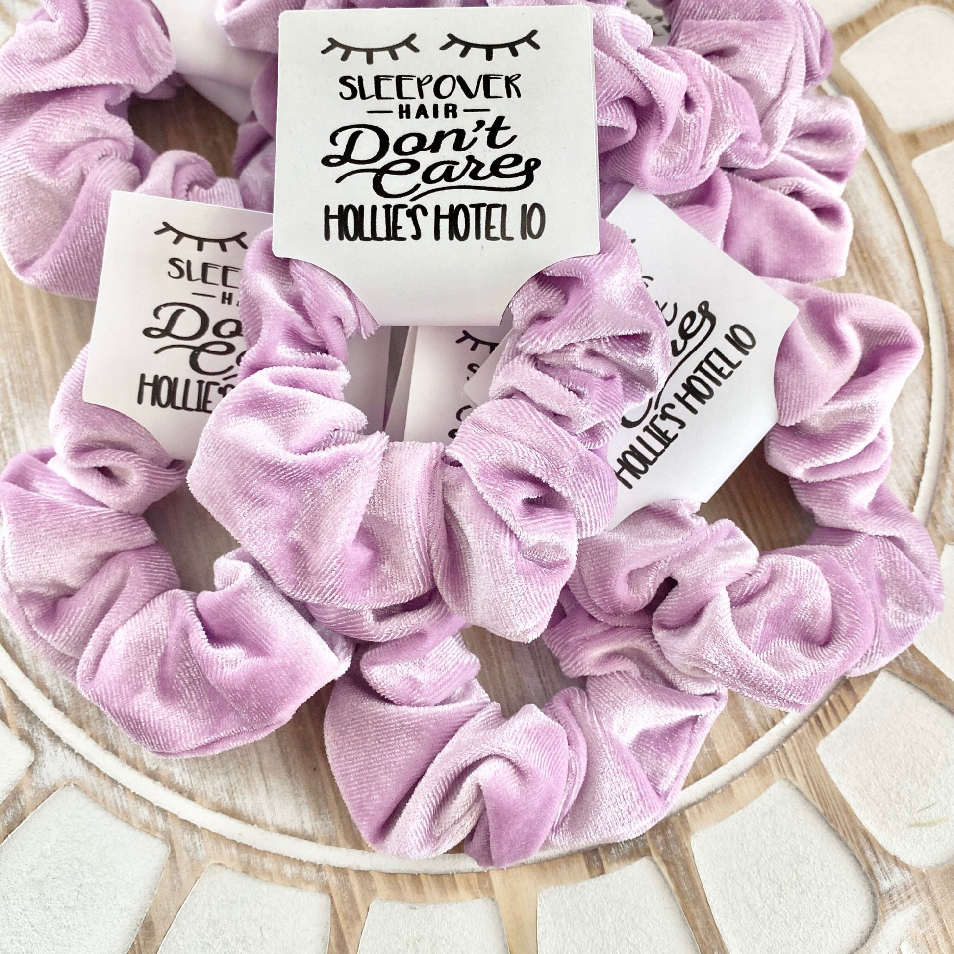 Lavendar scrunchies with 'Sleepover Hair Don't Care Hollie's Hotel 10' labels on a wooden surface.