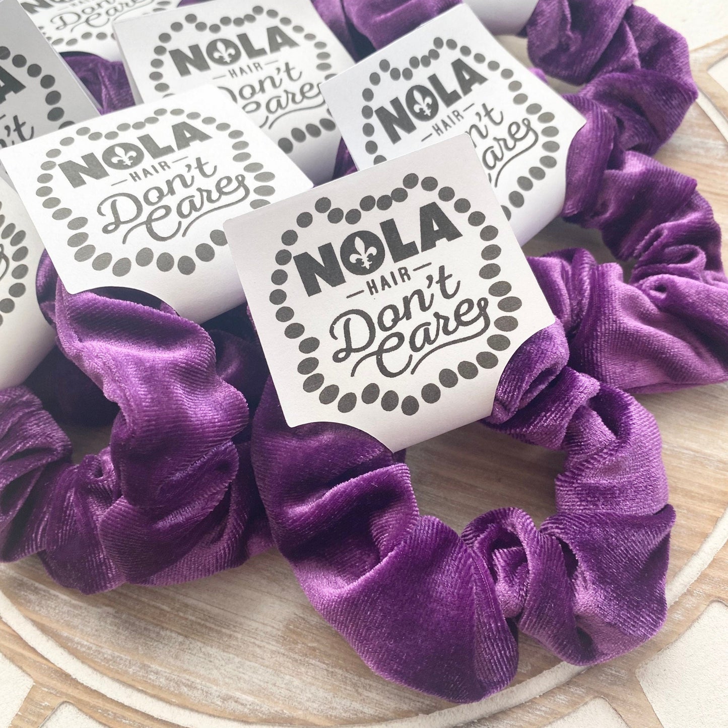 Purple scrunchies with 'NOLA Hair Don't Care' labels on a wooden surface