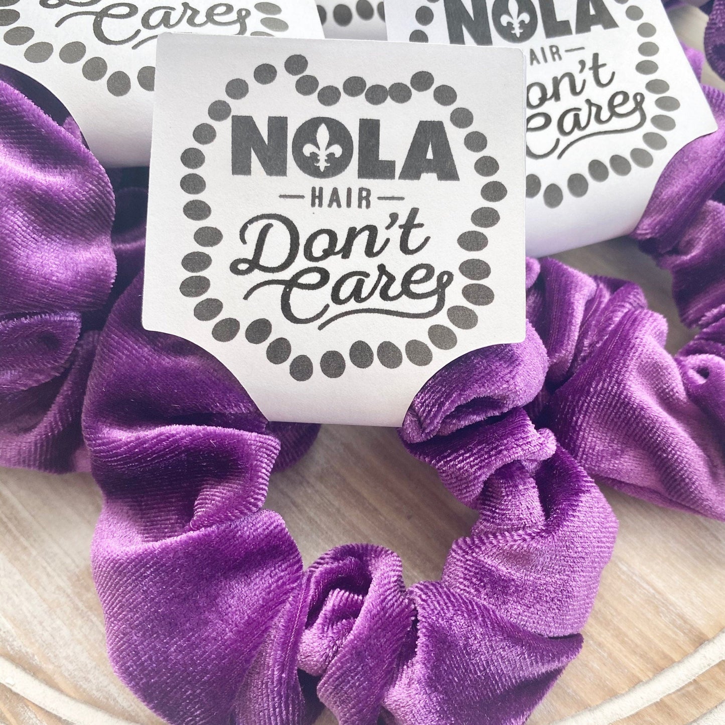 Purple scrunchies with 'NOLA Hair Don't Care' stickers on a wooden surface