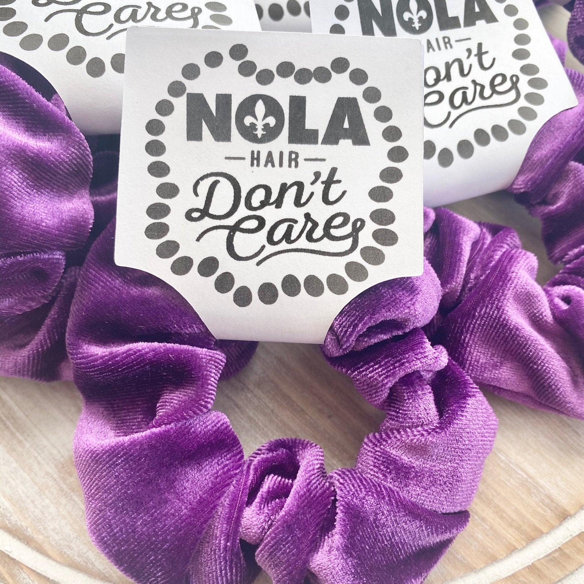 Purple scrunchies with 'NOLA Hair Don't Care' stickers on a wooden surface