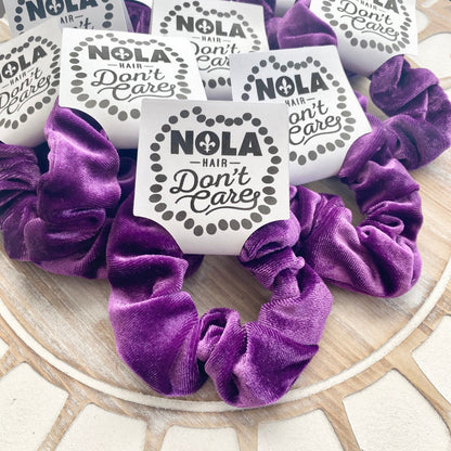 Purple scrunchies with 'NOLA Hair Don't Care' labels on a wooden surface
