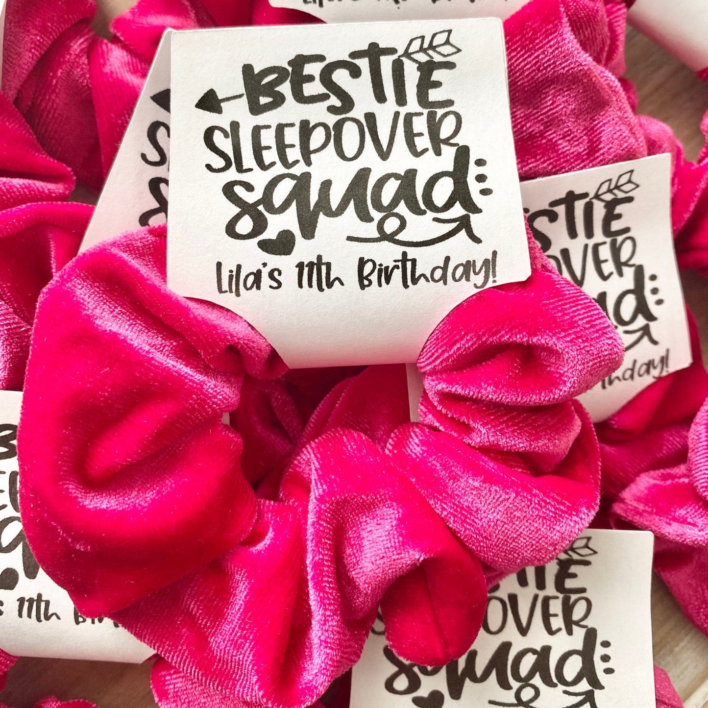Pink scrunchies with 'Bestie Sleepover Squad' labels on a wooden surface
