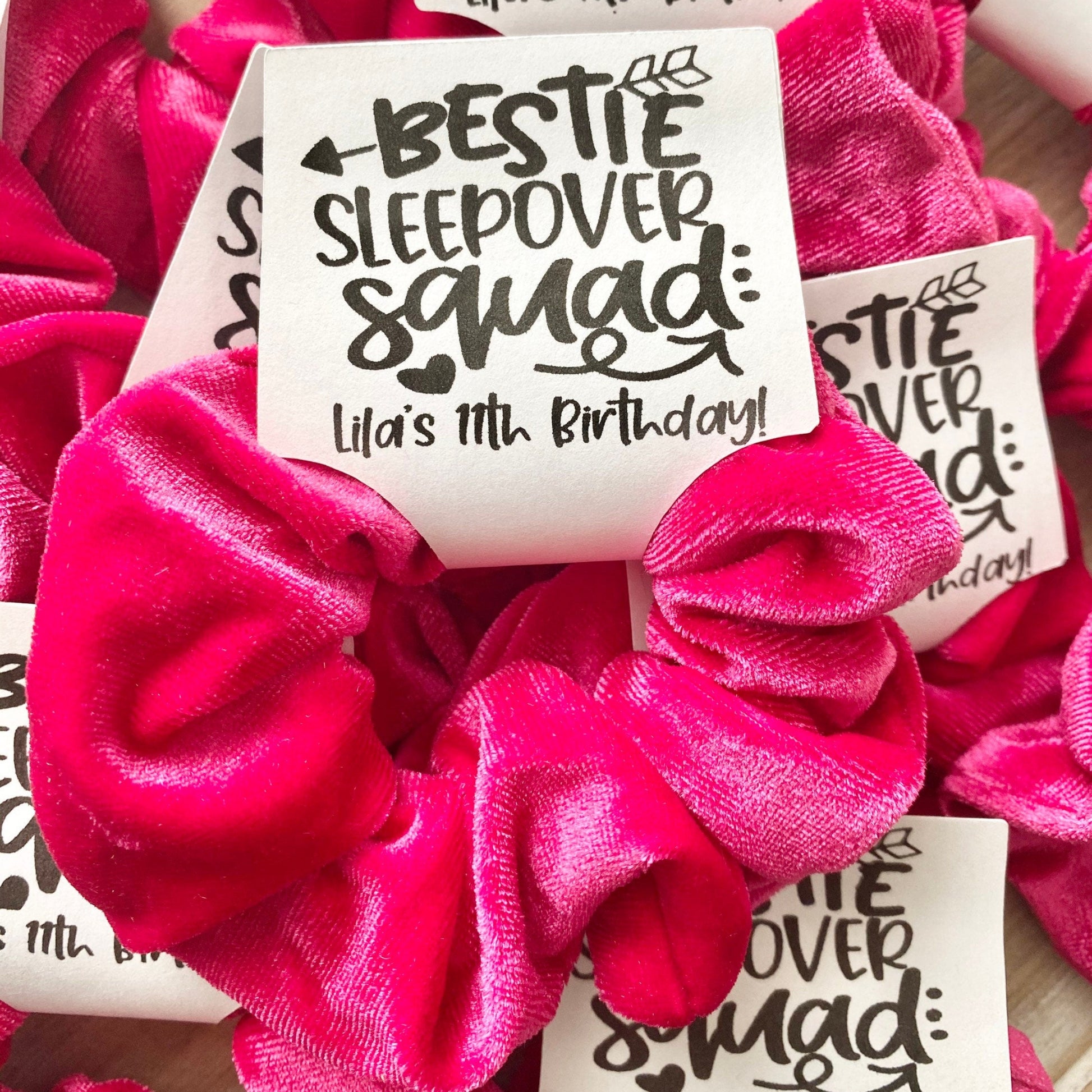 Pink scrunchies with 'Bestie Sleepover Squad' labels on a wooden surface