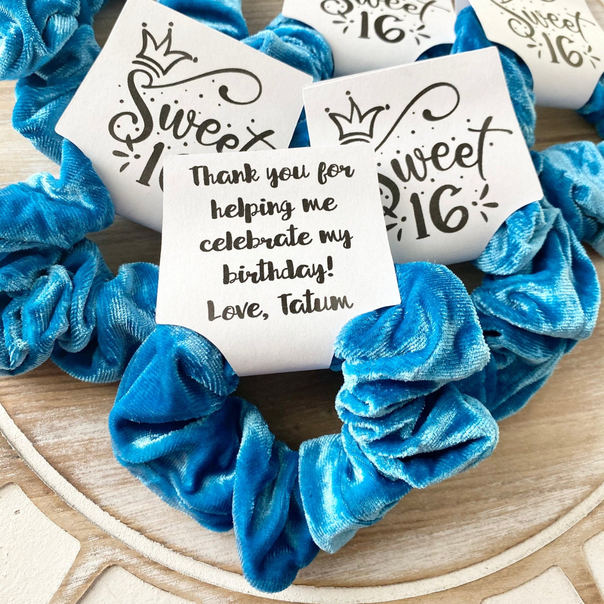 Blue scrunchies with personalized 'Sweet 16' cards on a wooden surface