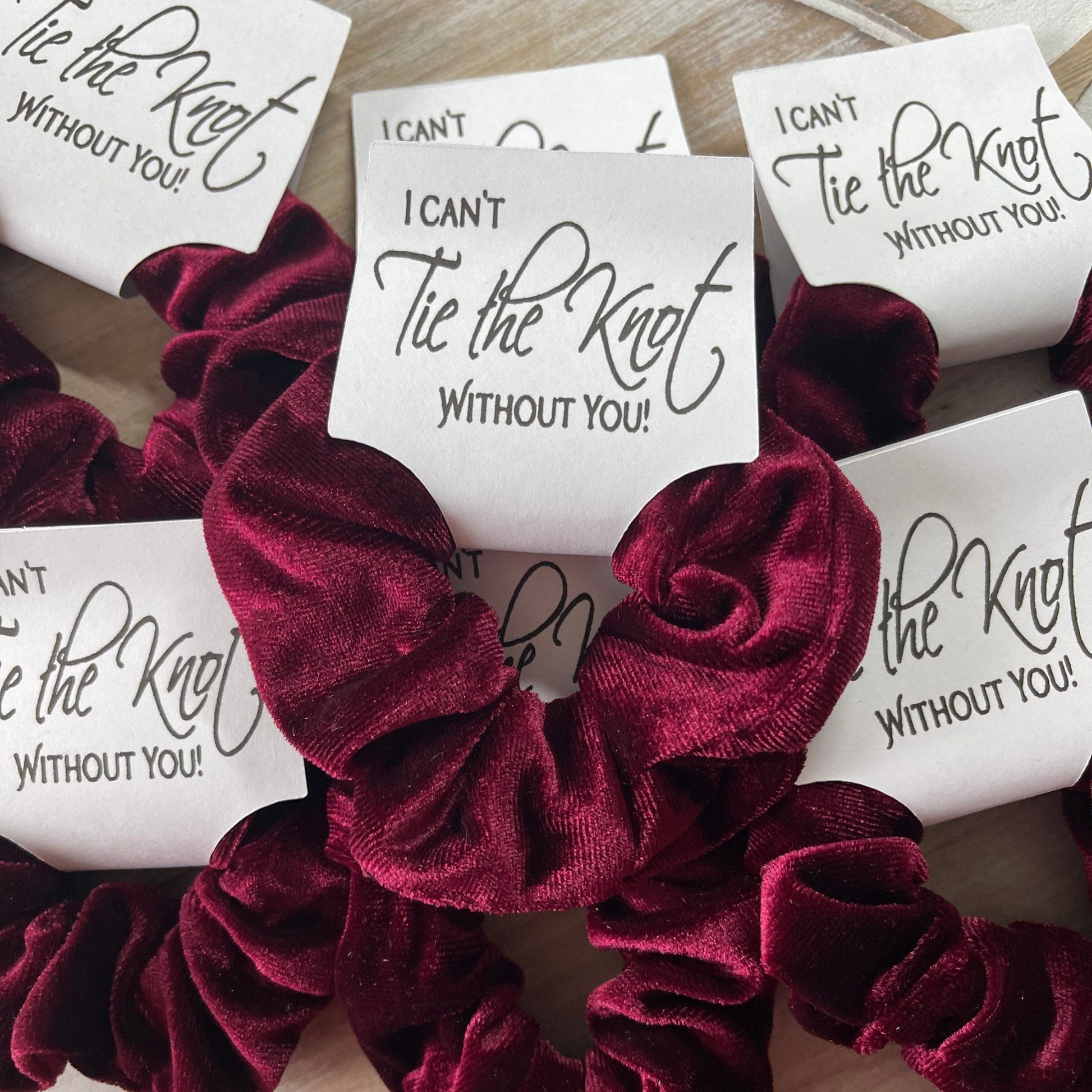 Maroon velvet scrunchies with 'I Can't Tie the Knot Without You' cards on a wooden surface