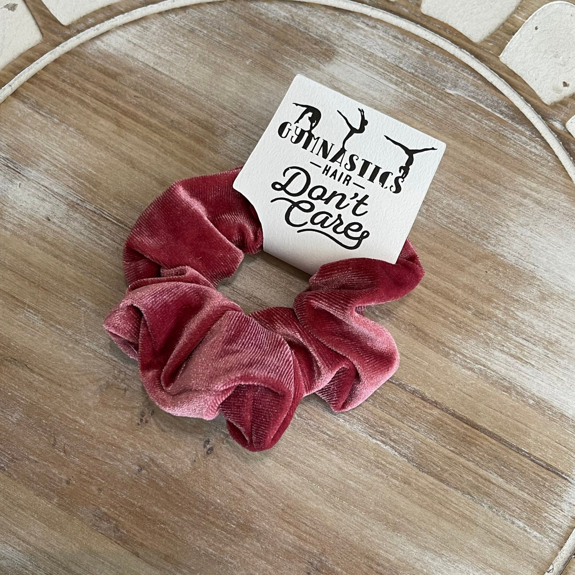 Pink scrunchie with a card reading 'Gymnastics Hair Don't Care' on a wooden surface