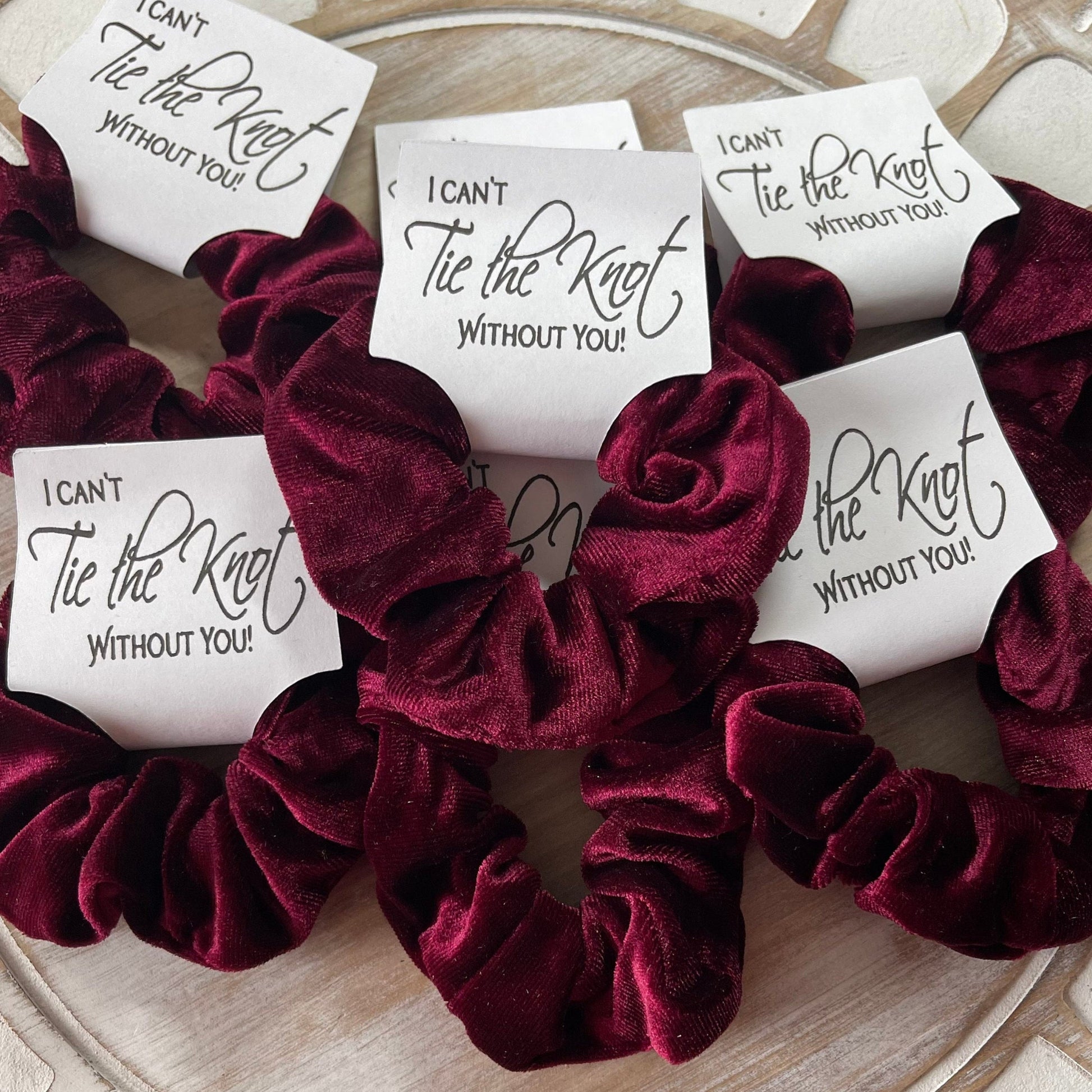 Maroon velvet scrunchies with 'I can't tie the knot without you' cards on a wooden surface.