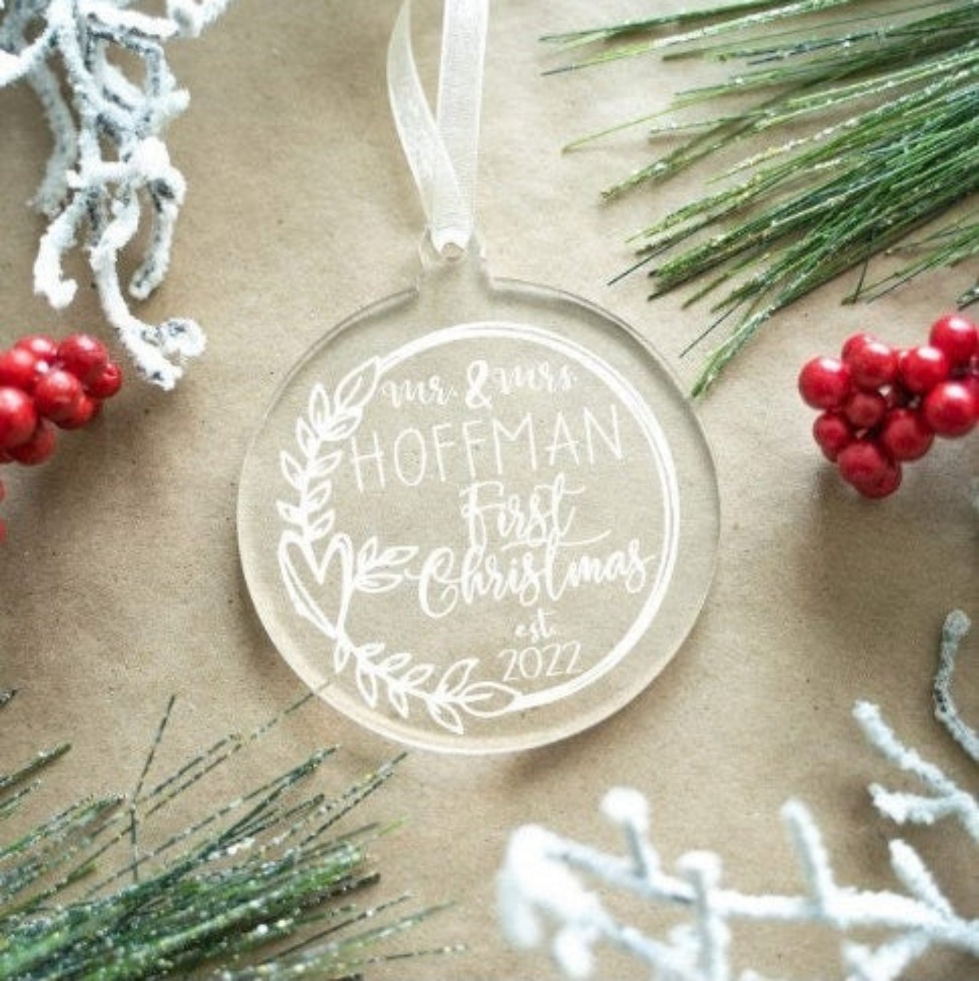 Clear acrylic ornament with white text surrounded by Christmas decorations on a beige background