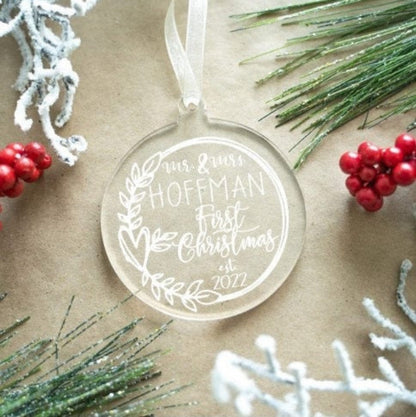 Clear acrylic ornament with white text surrounded by Christmas decorations on a beige background