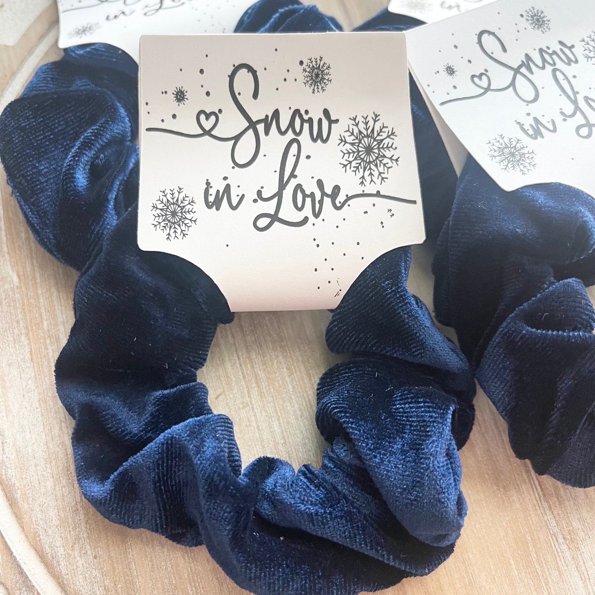 Navy blue scrunchies with 'Snow in Love' tags on a wooden surface