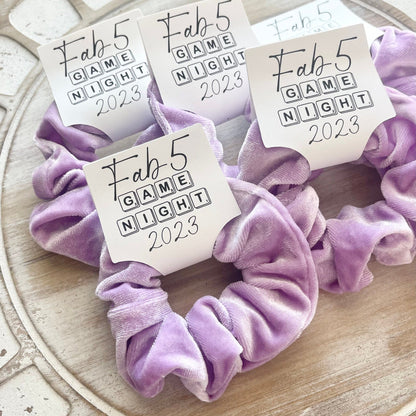 Purple scrunchies with 'Fab 5 Game Night 2023' labels on a decorative surface.