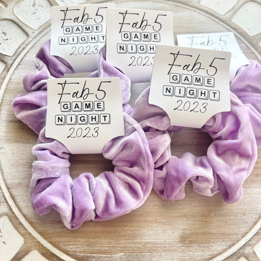 Two purple scrunchies with 'Fab 5 Game Night 2023' labels on a decorative surface.
