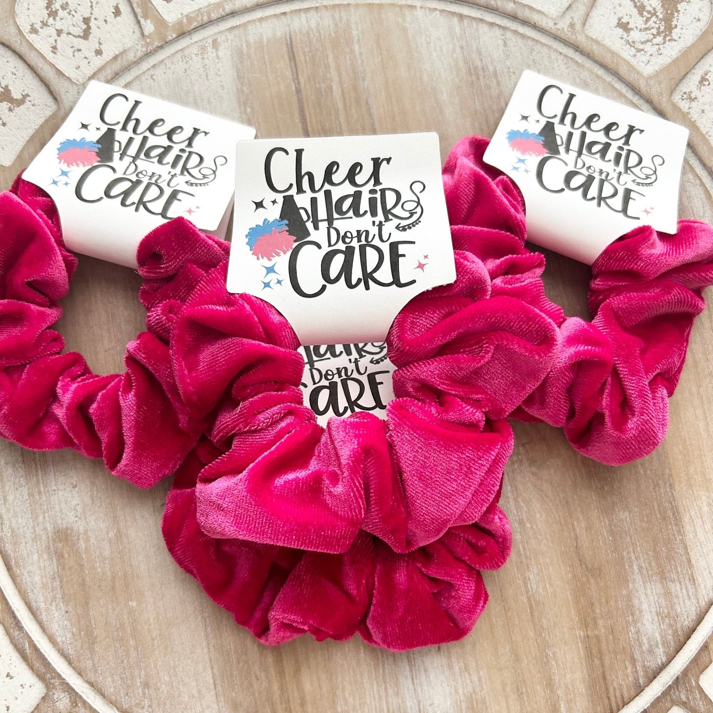 Pink scrunchies with 'Cheer Hair Don't Care' tags on a decorative wooden surface