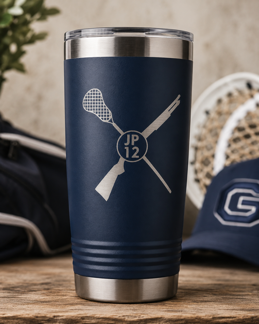 A navy blue insulated travel tumbler with stainless steel accents, featuring a white laser-engraved crossed lacrosse theme with a circular 'JP 12' logo. The tumbler is shown on a wooden table with a blurred navy lacrosse cap and gear in the background.