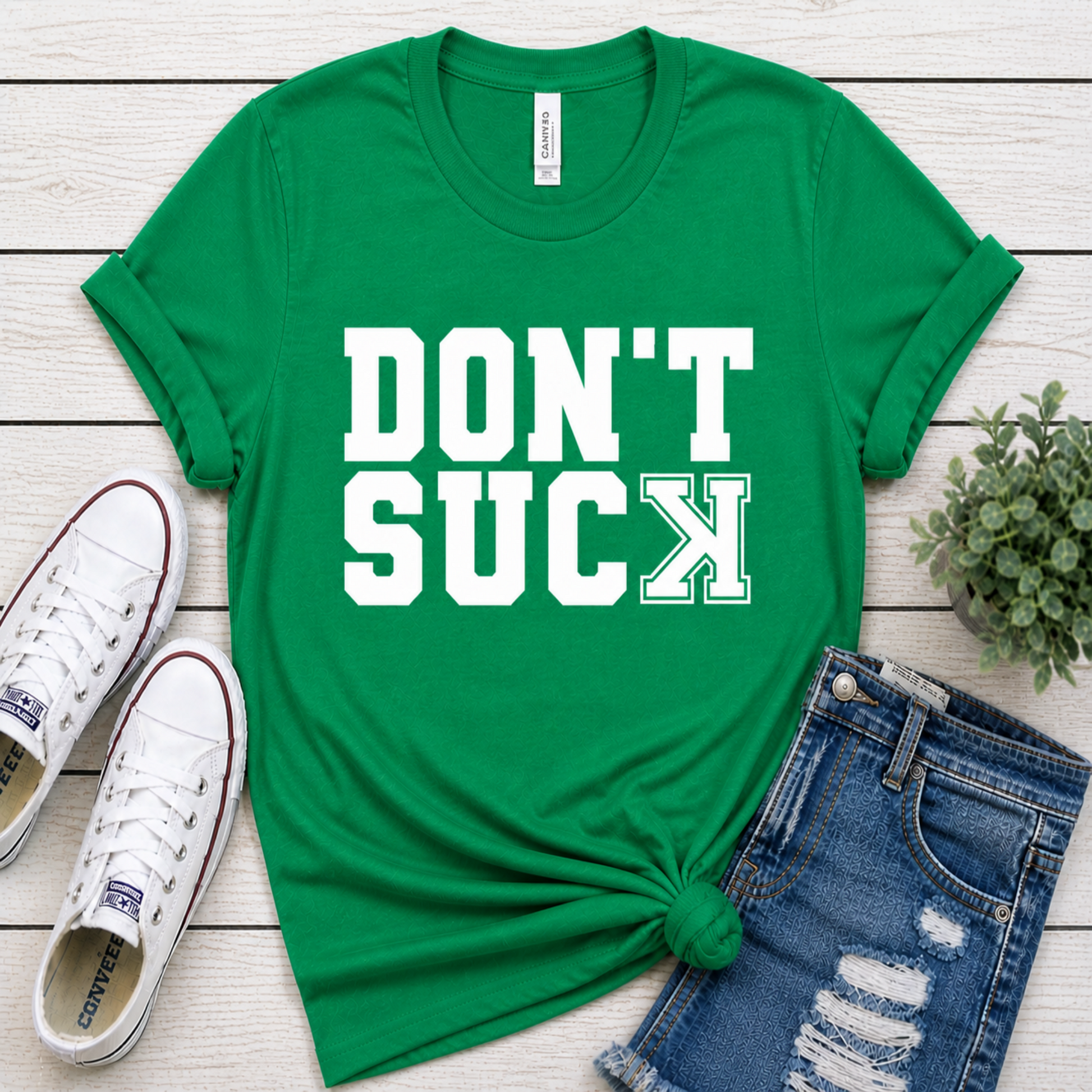 Green t-shirt with 'DON'T SUCK' text on a wooden surface with shoes and shorts.