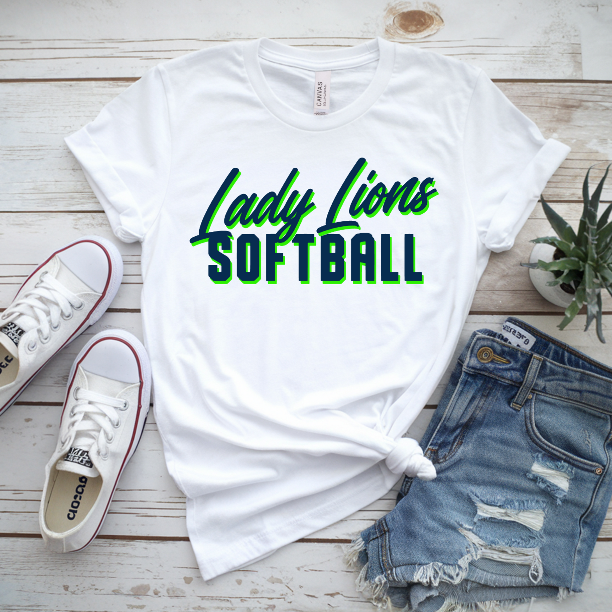 White t-shirt with 'Lady Lions Softball' text, Adidas sneakers, denim shorts, and a plant on a wooden surface.