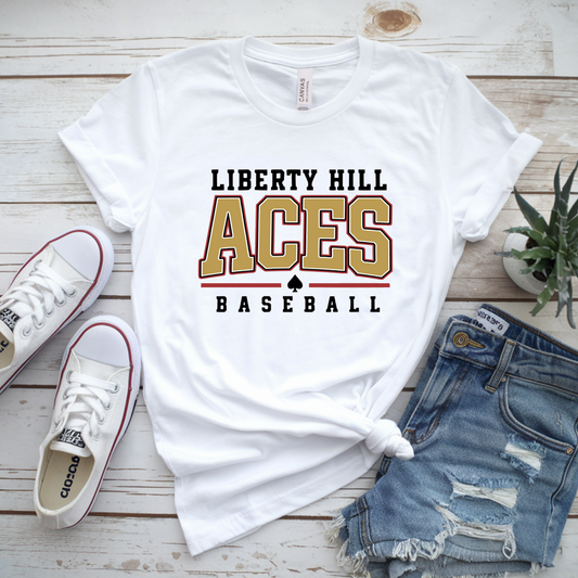 White graphic T-shirt laid flat on a whitewashed wood surface, styled with white Converse sneakers, distressed denim shorts, and a small potted plant. The shirt reads 'LIBERTY HILL ACES BASEBALL' with a large gold-and-black 'ACES' and a black spade graphic.