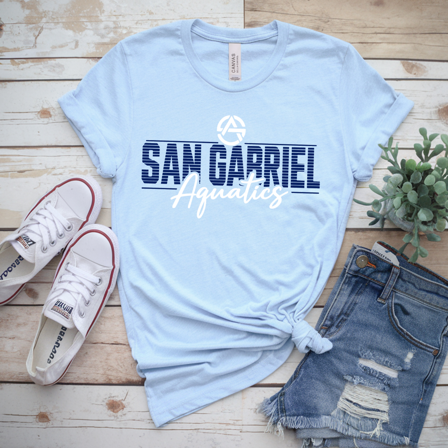 Light blue t-shirt with 'San Gabriel Aquatics' text, white sneakers, denim shorts, and a plant on a wooden surface.