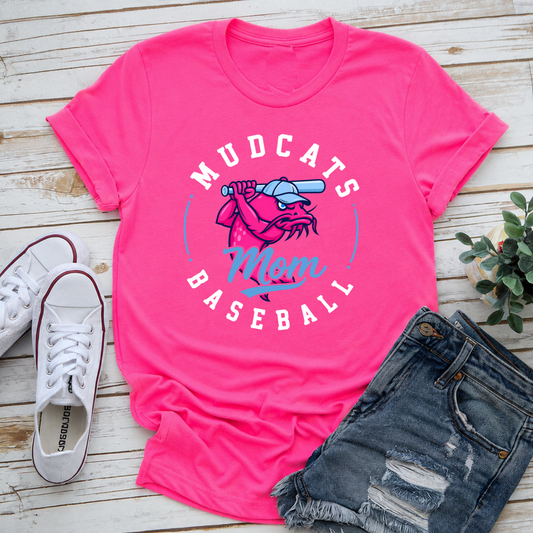 A bright pink graphic t-shirt laid flat on a light wood surface, styled with white sneakers, distressed denim shorts, and a small green plant. The shirt features a circular 'Mudcats Mom Baseball' graphic with a pink and blue cat holding a baseball bat.