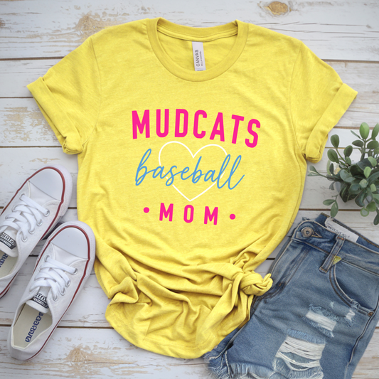 A bright yellow graphic T-shirt laid flat on a whitewashed wood surface, styled with white sneakers, distressed denim shorts, and a small green plant. The shirt reads “MUDCATS baseball MOM” in pink and blue lettering with a white heart outline.