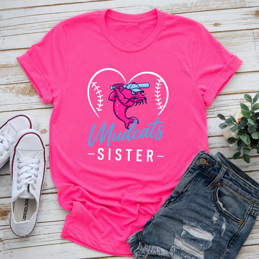 A bright pink graphic T-shirt laid flat on a light wood surface, styled with white sneakers, distressed denim shorts, and a small green plant. The shirt graphic shows a baseball-themed mascot inside a heart outline with the words 'Madcats SISTER'.