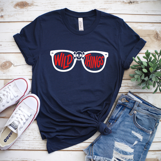 Navy blue t-shirt with 'Wild Things' sunglasses graphic, white sneakers, and denim shorts on a wooden surface.