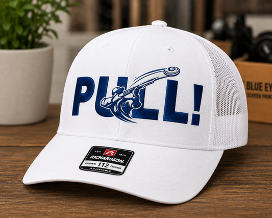 A white Richardson 112 trucker cap with a blue 'PULL!' graphic on the front, displayed on a wooden surface with a blurred indoor background.