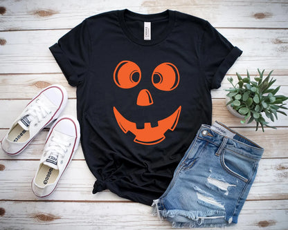 Black t-shirt with orange pumpkin face design, white sneakers, denim shorts, and a plant on a wooden surface.