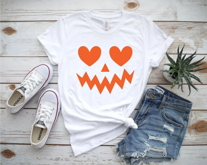 White t-shirt with orange heart and triangle pumpkin face design, paired with white sneakers and denim shorts.